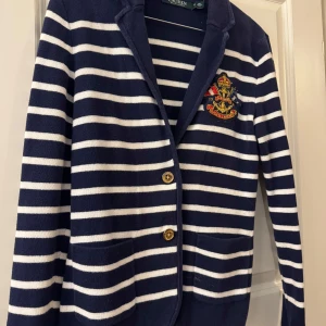 Randig cardigan jacka från Ralph Lauren - Snygg stickad cardigan jacka från Ralph Lauren i marinblått och vitt med horisontella ränder. Jackan har två guldknappar, fickor framtill och en broderad patch på bröstet. Klassisk krage och långärmad modell, perfekt för en preppy look.