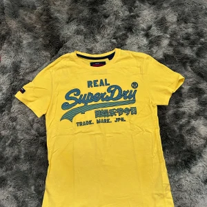 Gul Superdry t-shirt med tryck - Gul t-shirt från Superdry med blått, randigt logotryck och japanska tecken på bröstet. Klassisk rund hals och korta ärmar. Tillverkad i mjuk bomull, perfekt för en avslappnad stil.