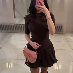 Brun kort klänning med volang - Använd 1-2 ggr. Zara. Supersnygg brun klänning från Zara med kort volangkjol och ribbad överdel. Klänningen har rund hals, halvlånga ärmar och en loose passform över kjolen. Perfekt för dig som gillar stilrena och trendiga plagg med lite edge.