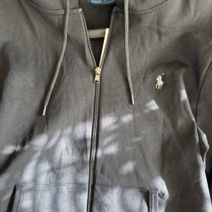 Svart hoodie från Polo Ralph Lauren - Svart hoodie från Polo Ralph Lauren med dragkedja, huva med snörning och klassisk broderad logga på bröstet. Tillverkad i mjuk bomull och har praktiska fickor framtill. Perfekt för en chill och stilren look. Helt ny, aldrig använd.
