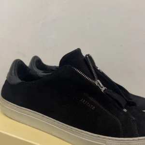 Svarta sneakers från Axel Arigato - Svarta sneakers från Axel Arigato i mocka med vit sula och cool dragkedjedetalj framtill istället för klassisk snörning. Snygg minimalistisk design med diskret logga på sidan. Perfekta för dig som gillar stilrena och moderna skor.