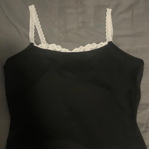 Svart linne med spets från SHEIN - Svart linne från SHEIN i storlek XS med vita spetsdetaljer längs axelbanden och urringningen. Axelbanden är smala och dekorerade med blommig spets som ger en söt kontrast till det svarta tyget. Perfekt att styla med jeans eller kjol.
