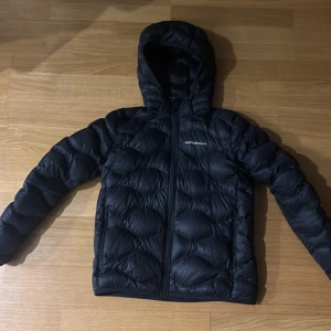 Svart dunjacka Peak Performance 160 - Svart dunjacka från Peak Performance med huva och broderad logga på bröstet. Jackan har quiltad design, dragkedja framtill och lång ärm. Perfekt för kyliga dagar och riktigt snygg streetstil. Jackan är i jättebra skick och har inga defekter 