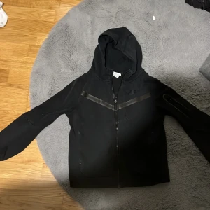 Svart Nike hoodie med dragkedja - Svart hoodie från Nike med hel dragkedja framtill och huva. Snygga detaljer med svarta band över bröstet och en praktisk ficka med dragkedja på ena ärmen. Perfekt för chill eller träning, stilren och enkel design.