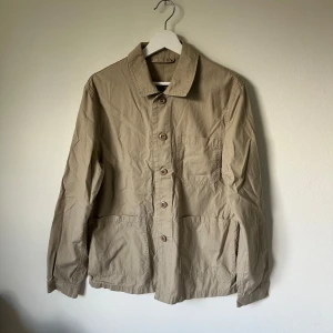 Beige overshirt från Mango Man - Stilren beige overshirt från Mango Man i 100% bomull. Jackan har klassisk krage, knäppning med bruna knappar framtill och tre praktiska fickor. Perfekt för lager-på-lager och enkel att matcha med det mesta. 2H6