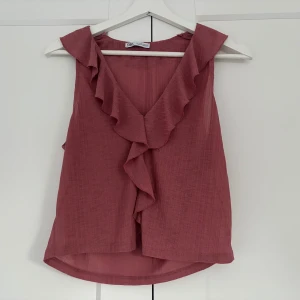 Rosa volangblus från Zara - Säljer en söt ärmlös blus från Zara i en dämpad rosa nyans. Toppen har v-ringning med volangdetaljer längs halsringningen och framtill. Lätt och luftig i ett tunt tyg, perfekt för varmare dagar.