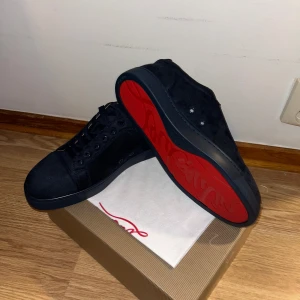 Svarta sneakers från Christian Louboutin - Snygga svarta sneakers från Christian Louboutin i mocka med klassisk röd sula och broderad logga på hälen. Skorna har rund tå, snörning och beige innersida. Perfekta för dig som vill sticka ut med en lyxig touch.