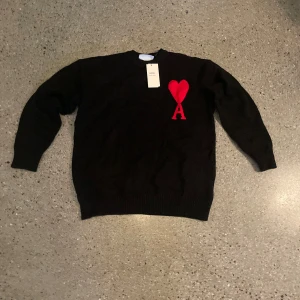 Svart stickad tröja från Ami - Svart stickad tröja från Ami Alexandre Mattiussi med rött hjärta och bokstaven A broderat på bröstet. Klassisk rund halsringning och långa ärmar. Perfekt för dig som vill ha en stilren men ändå unik look.