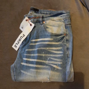 Blå jeans från Purple Brand, stl 38 - Säljer ett par blå jeans från Purple Brand i storlek 38. Jeansen har en cool blekt tvätt med vita stänk och ränder framtill, samt klassiska femficksdetaljer. Materialet är jeans och passformen är rak med normal midja. Perfekt för dig som gillar streetwear med edge.