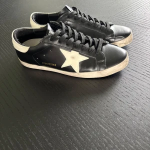 Golden Goose svart/vita sneakers - Snygga Golden Goose sneakers i svart skinn med vit stjärndetalj på sidan och vit häl. Klassisk låg modell med svarta skosnören och GGDBISSTAR-tryck i guld. Skorna har platt sula och rund tå.