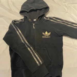Svart Adidas hoodie med guldtryck - Svart hoodie från Adidas med ikoniska guldiga ränder längs ärmarna och stort guldigt Adidas-tryck både fram och bak. Tröjan har huva, dragkedja och känguruficka. Perfekt för dig som gillar sportig och street-inspirerad stil. PRIS KAN DISKUTERAS!