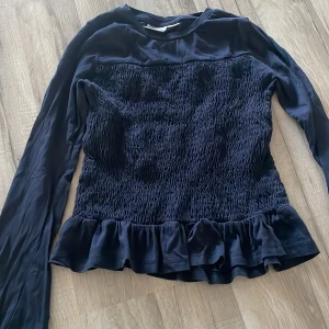 Mörkblå volangblus från Gina Tricot - Supersöt mörkblå blus från Gina Tricot med långa ärmar, smockad kropp och volangkant nedtill. Blusen har en mjuk, glansig känsla upptill och är perfekt för dig som gillar trendiga detaljer och en feminin look.