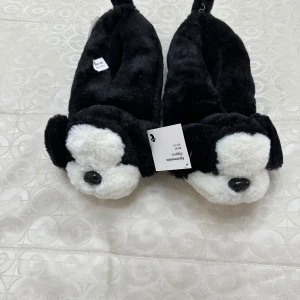 Svarta och vita tofflor med hundmotiv - Mysiga tofflor i svart och vitt med gulligt hundansikte framtill. Tillverkade i mjuk plysch som håller fötterna varma och sköna. 