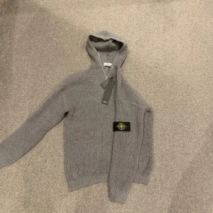 Grå stickad hoodie från Stone Island - Passar både L och M……. Säljer en grå stickad hoodie från Stone Island med klassisk logga på ärmen. Tröjan har huva, half zip-dragkedja och är långärmad. Perfekt för dig som gillar streetwear och vill ha något både snyggt och bekvämt. Materialet är mjukt och värmande ull.