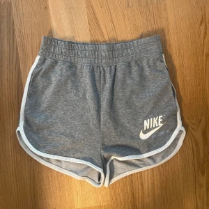 Grå Nike Sportswear shorts XS - Snygga grå Nike Sportswear shorts i mjuk bomull med vit kant och logga framtill. Modellen har elastisk midja och rundade benkanter för en sportig vibe. Perfekta för chill eller träning, riktigt sköna och stilrena.