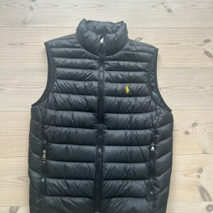 Svart dunväst från Polo Ralph Lauren XS - Svart dunväst från Polo Ralph Lauren med gul broderad logga på bröstet. Västen har quiltad design, två dragkedjefickor och hel dragkedja framtill. Insidan har ett mörkgrönt och marinblått rutigt mönster. Västen är i mycket gott skick utan hål och fläckar. Observera även ath västen är relativt stor i storlek!