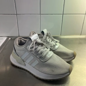 Vita Adidas sneakers  - Snygga vita Adidas sneakers med klassiska tre ränder på sidan. Ovandelen är i mesh och syntet, vilket gör dem lätta och luftiga. Skorna har snörning och en platt sula med gummidetaljer. Perfekta för dig som gillar stilrena och sportiga skor. ( behövs tvättas ) 