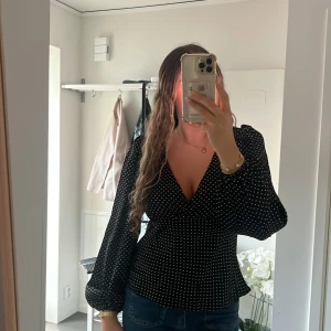 Svart prickig blus från Neo Noir - Supersnygg svart blus från Neo Noir med vita prickar, v-ringning och långa puffiga ärmar. Blusen har en figurnära passform i midjan och är tillverkad i ett lätt och mjukt material som känns skönt mot huden. Perfekt att styla med jeans eller kjol.