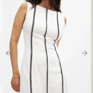 Vit långklänning med svarta detaljer - Vit långklänning med svarta dekorativa sömmar i lodräta rader. Klänningen är ärmlös, har öppen rygg med knytband och en smickrande, figurnära passform. Perfekt för dig som vill sticka ut med en stilren och modern look. Helt oanvänd med etiketten kvar på. Säljer pga fel stl då jag inte hann returnera i tid. Inköpt för 800kr, mitt pris 500kr