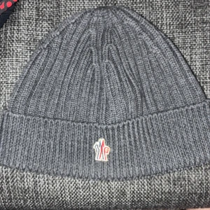 Grå ribbad mössa från Moncler - Snygg grå ribbstickad mössa från Moncler Grenoble med uppvikt kant och diskret logotyp framtill. Tillverkad i mjuk ull som håller dig varm och ger en clean look. Perfekt för dig som vill ha en stilren accessoar med exklusiv känsla.