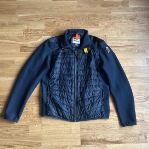 Mörkblå quiltad jacka Parajumpers M - Snygg mörkblå jacka från Parajumpers i storlek M. Jackan har quiltad front och rygg, släta ärmar och coola detaljer som gul rem vid dragkedjan. Materialmix med polyester och syntet, perfekt för kyliga dagar. Klassisk ståkrage och hel dragkedja framtill.