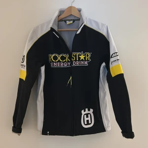 Rockstar x Husqvarna svart/vit jacka - Snygg svart och vit jacka från Husqvarna och Rockstar Energy Drink med gula detaljer och tryck på bröst, rygg och ärmar. Jackan har dragkedja framtill, hög krage och sportig look. Perfekt för dig som gillar motorsport och vill sticka ut.