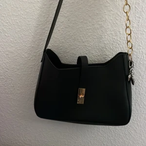 Svart axelväska med guldkedja - Stilren svart axelväska i syntetmaterial med guldfärgad kedja och låsdetalj framtill. Väskan har en minimalistisk design med raka linjer och en smidig axelrem. Perfekt för dig som gillar en clean look med en touch av guld.