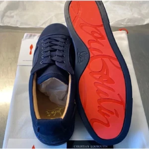 Christian Louboutin marinblå sneakers - Snygga marinblå sneakers från Christian Louboutin med klassisk röd sula och diskret logga. Skorna har rund tå, snörning och är tillverkade i mocka och textil. Perfekta för dig som vill sticka ut med exklusiv stil och detaljer.