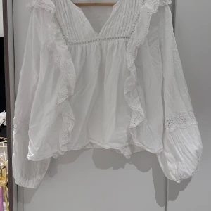 Vit blus med volang från Gina Tricot - Romantisk vit blus från Gina Tricot med v-ringning, volangdetaljer och broderade spetsinslag. Långa puffärmar med resår vid ärmslut och dekorativa spetsband över ärmarna. Perfekt för dig som gillar en feminin och luftig stil.