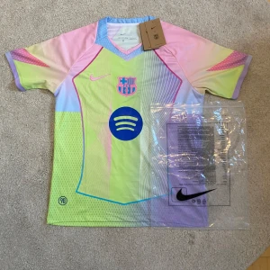 FC Barcelona x Nike pastell fotbollströja - Snygg FC Barcelona fotbollströja från Nike i pastellfärger med klubbmärke och Spotify-logga på bröstet. Tröjan har korta ärmar, V-ringning och är tillverkad i lätt Dri-FIT material som andas. Färgerna är limegrön, rosa, ljusblå och lila med grafiska linjer.
