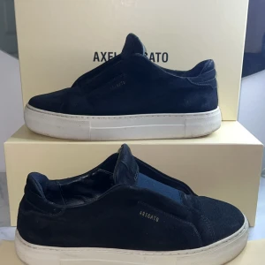 Mörkblå sneakers från Axel Arigato - Snygga mörkblå sneakers från Axel Arigato i mocka med vit platt sula. Minimalistisk design utan snörning, elastiskt band över vristen och diskret logga på sidan. Perfekta för dig som gillar stilrena och moderna skor. Vid frågor, tveka inte att kontakta mig! 