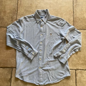 Ljusblå skjorta från Ralph Lauren - Snygg ljusblå skjorta från Ralph Lauren i slim fit-modell. Klassisk button down-krage, broderad logga på bröstet och vita knappar. Tillverkad i bomull med en stickad oxfordstruktur. Perfekt för dig som gillar stilrena och tidlösa plagg.krympte lite i tvätten