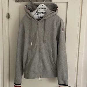 Grå Moncler hoodie med dragkedja - Snygg grå hoodie från Moncler med hel dragkedja, huva och snörning. Tröjan har Moncler-logga på ärmen och med röd, vit och marinblå rand vid handlederna och på luvan. perfekt för chill dagar och för fin dagar. Fickor framtill och klassisk passform. Riktigt allmänt skön tröja. Nypris ca 5000. Mitt pris är inte hugget i sten