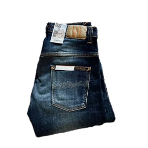 Nudie Jeans Thin Finn Indigo Dreams W30 L34 - Snygga blå jeans från Nudie Jeans, modellen Thin Finn Indigo Dreams. Klassisk femficksdesign med orangea sömmar och Nudie-logga på läderpatchen bak. Jeansen har en smal passform och är tillverkade i mjukt denimtyg. Perfekt för dig som gillar stilrena och moderna jeans.