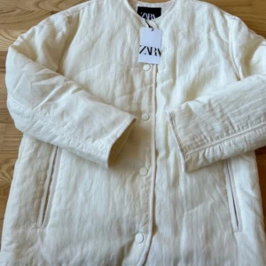 Vit quiltad jacka från Zara - Snygg vit quiltad jacka från Zara med rund halsringning och tryckknappar framtill. Jackan har raka ärmar och två fickor på sidorna. Perfekt för dig som gillar minimalistisk och clean stil.