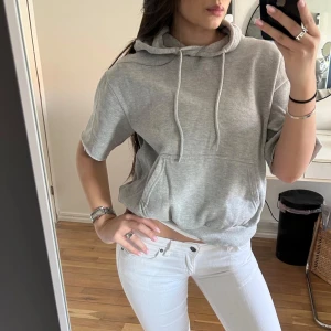 Grå croppad hoodie med kort ärm - Säljer en grå croppad hoodie med korta ärmar och snörning i huvan. Tröjan har en stor magficka framtill och är gjord i mjuk bomullsmix. Perfekt för chill dagar och enkel att matcha med jeans eller leggings.