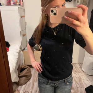 Svart t-shirt med små prickar - Säljer en mörkblå stickad t-shirt med små guldiga prickar. T-shirten har en klassisk rund hals och korta ärmar. Perfekt för en avslappnad stil.