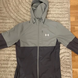 Grå och svart vindjacka Under Armour - Snygg tvåfärgad vindjacka från Under Armour i grått och svart. Jackan har huva, hel dragkedja framtill och en bröstficka med dragkedja. Perfekt för blåsiga dagar och sportiga outfits. Under Armour-logga på bröstet ger en sportig vibe.