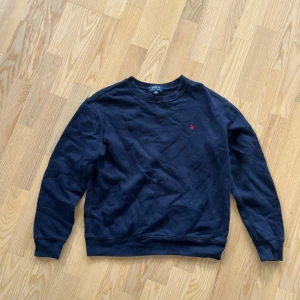 Mörkblå sweatshirt från Polo Ralph Lauren - Klassisk mörkblå sweatshirt från Polo Ralph Lauren med rund halsringning och röd broderad logga på bröstet. Tröjan har långa ärmar och ribbade muddar vid ärmslut och nederkant. Perfekt för en chill och stilren look.
