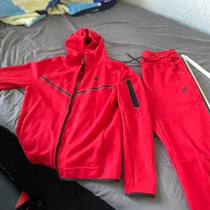 Röd Nike hoodie med dragkedja S - Nike hoodie i starkt röd färg med hel dragkedja, huva och svarta detaljer. Tröjan har en ficka med dragkedja på ärmen och svart Nike-logga på bröstet. Tillverkad i mjuk fleece, perfekt för chill eller träning.