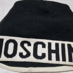 Svart-vit mössa från Moschino - Snygg svart stickad mössa från Moschino med bred vit kant och stor svart logotyp runt om. Perfekt accessoar för att sticka ut i vinterkylan och visa din stil med ett ikoniskt märke.