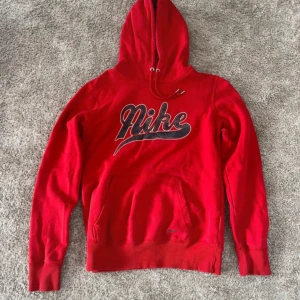 Röd Nike hoodie med broderad logga - Riktigt snygg röd hoodie från Nike med stor broderad logga i svart och vitt på bröstet. Tröjan har huva med snörning, känguruficka och är tillverkad i mjuk bomullsmix. Den är liten i storleken och skulle kunna vara en s.
