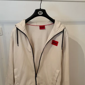 Beige hoodie från Hugo med dragkedja - Snygg beige hoodie från Hugo med svart dragkedja och svarta snören i huvan. Tröjan har röda Hugo-loggor på bröstet och insidan av nacken. Perfekt för en chill och stilren look. PRIS KAN DISKUTERAS 