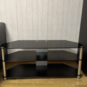 TV-bänk 500kr - Denna produkt är ett svart TV-bord eller mediabänk i glas och metall, med tre hyllplan och modern design. Perfekt för TV, spelkonsoler eller annan elektronik. Bordet ser ut att vara i mycket gott skick utan synliga repor eller skador. Passar utmärkt i vardagsrummet.