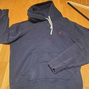 Marinblå hoodie från Polo Ralph Lauren - Marinblå hoodie från Polo Ralph Lauren med klassisk röd broderad logga på bröstet. Tröjan har huva med vit snörning, känguruficka och långa ärmar. Tillverkad i mjuk bomull, perfekt för chill dagar eller när du vill ha en clean streetstyle.