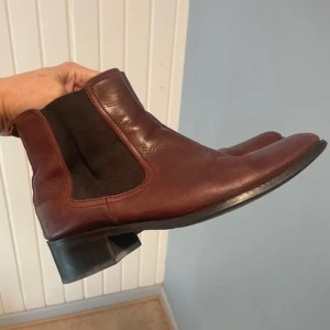 Bruna chelsea boots i skinn - Snygga bruna chelsea boots i skinn med svarta elastiska paneler på sidorna och låg klack. Klassisk rund tå och enkel att dra på tack vare hällan bak. Perfekta för dig som gillar stilrena och tidlösa skor.