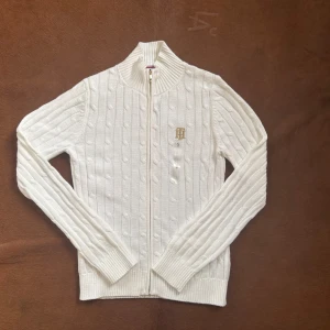 Vit kabelstickad kofta från Tommy Hilfiger - Kabelstickad vit kofta från Tommy Hilfiger med guldfärgad dragkedja och broderad logga på bröstet. Hög krage, ribbade muddar och hel dragkedja framtill. Perfekt för lager-på-lager och snygg till jeans eller kjol.