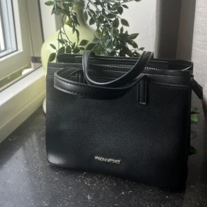 Svart handväska från Paco Martinez - Stilren svart handväska från Paco Martinez i skinnimitation med två handtag och flera fack, inklusive ett med dragkedja. Väskan har en klassisk, boxig form och silverfärgad logga framtill. Perfekt för dig som gillar minimalistisk och tidlös design. Går att fästa längre band.