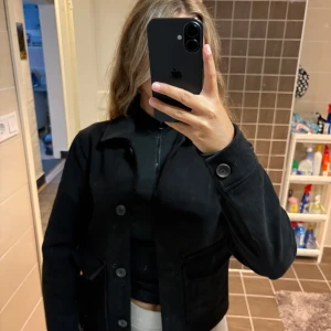 Svart mockajacka med knappar - Säljer en svart mockajacka MOCKAIMITATION!!! med klassisk krage och stora knappar framtill. Jackan har två fickor och långa ärmar med knappdetalj vid ärmslutet. Enkel och stilren modell som är lätt att matcha till olika outfits. Helt oanvänd köpte annan kapp. Nypris 600kr.