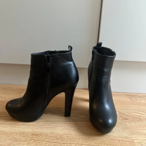 Svarta stövletter med klack - Snygga svarta stövletter från Attitude med hög klack och rund tå. Skorna har dragkedja i både guld och svart på sidorna och är gjorda i ett slätt material som liknar skinn. Perfekta för dig som vill ha en stilren och edgy look. Klacken är 12cm